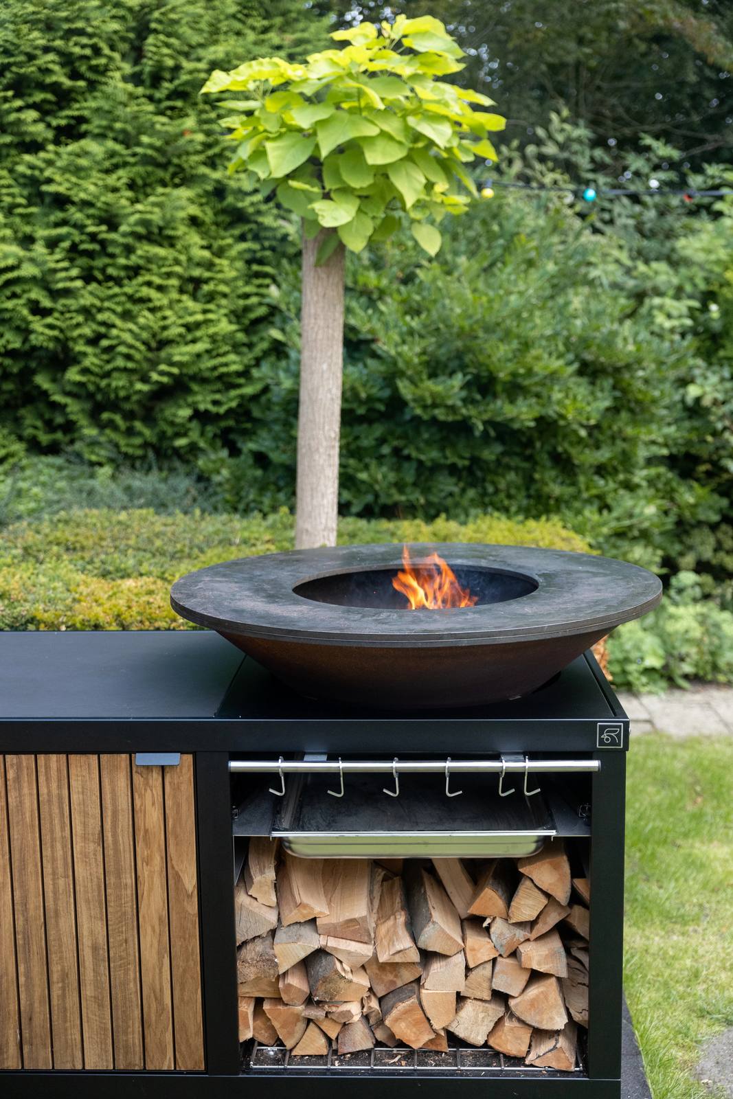 Zwarte OFYR buitenkeuken met brandende vuurschaal, houten deur en opslag voor brandhout in een groene tuin